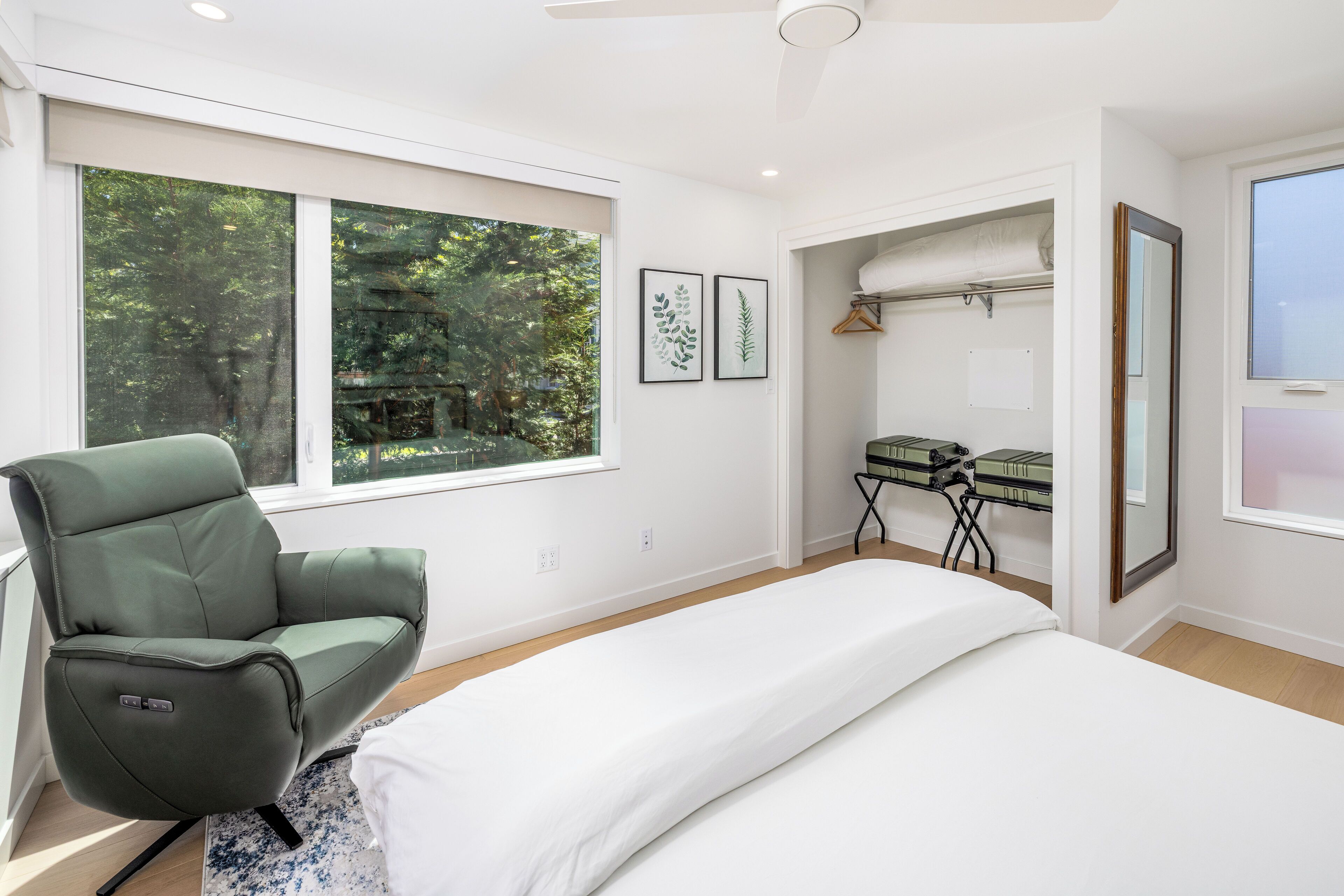 Une chambre moderne avec un fauteuil vert, un lit, un bureau avec une chaise et une grande fenêtre donnant sur des arbres.