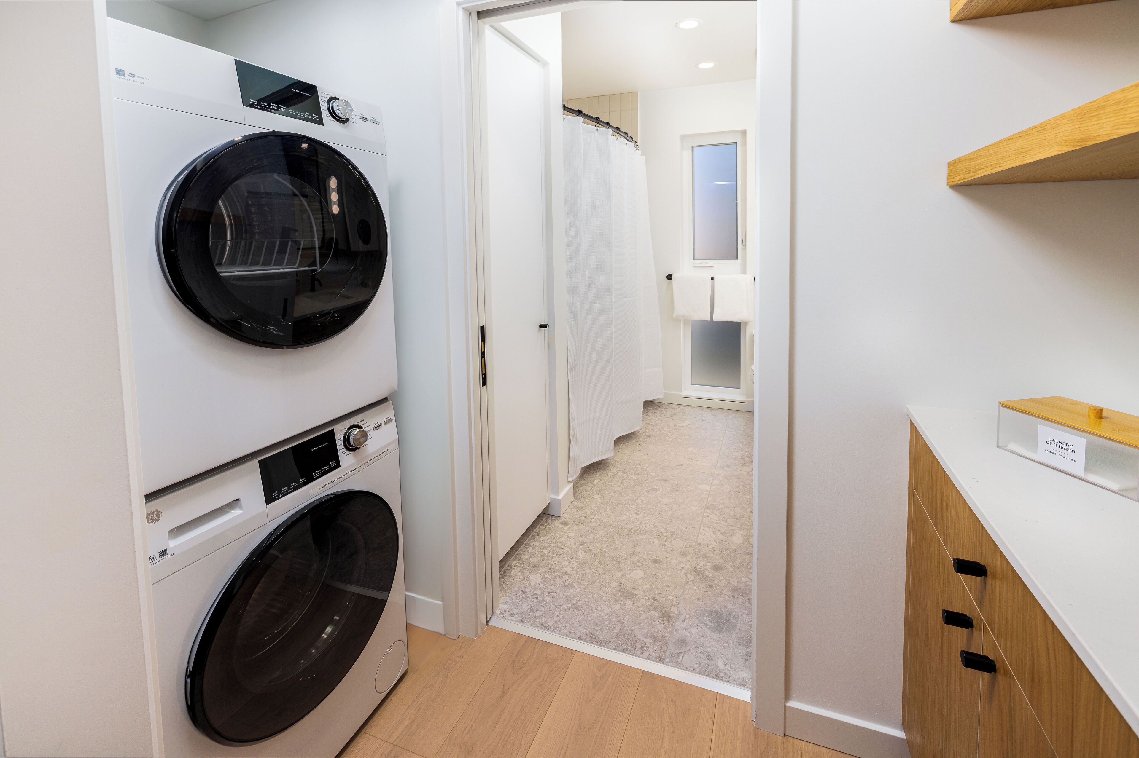 A laundry room with a front-loading washer and dryer, a wooden cabinet, and a shower area.