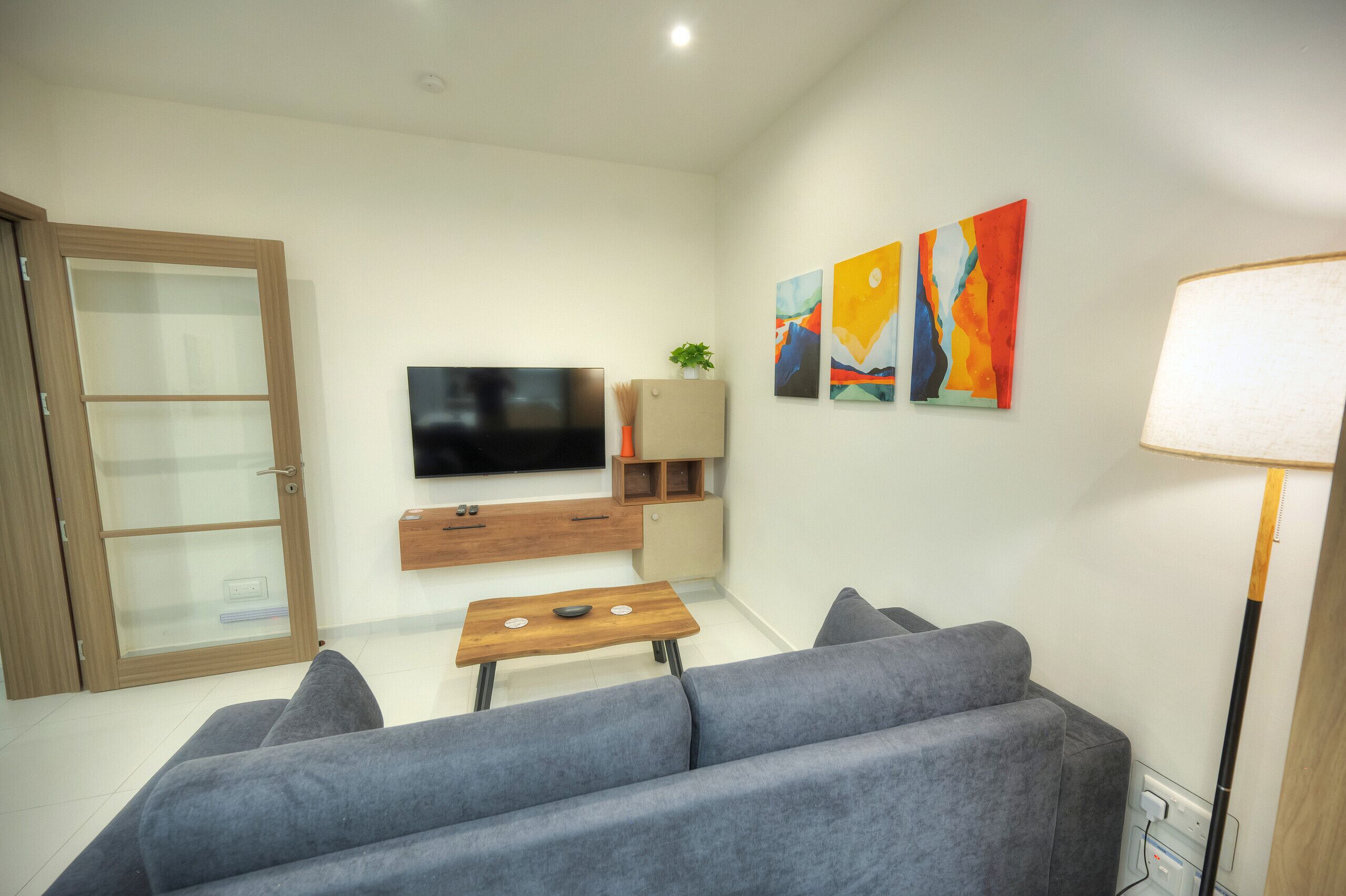 Uma sala de estar moderna com um sofá cinzento, mesa de centro de madeira e uma TV de tela plana fixada na parede. Há quadros abstratos na parede e um abajur.