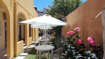 Un patio con mobiliario exterior, incluyendo mesas y sillas, bajo sombrillas, rodeado de follaje y flores.