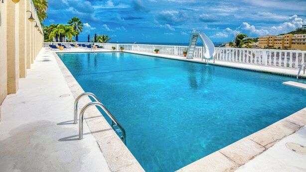 Une piscine d’un bleu éclatant, bordée d’une terrasse blanche, de chaises longues et d’une allée bordée de palmiers.