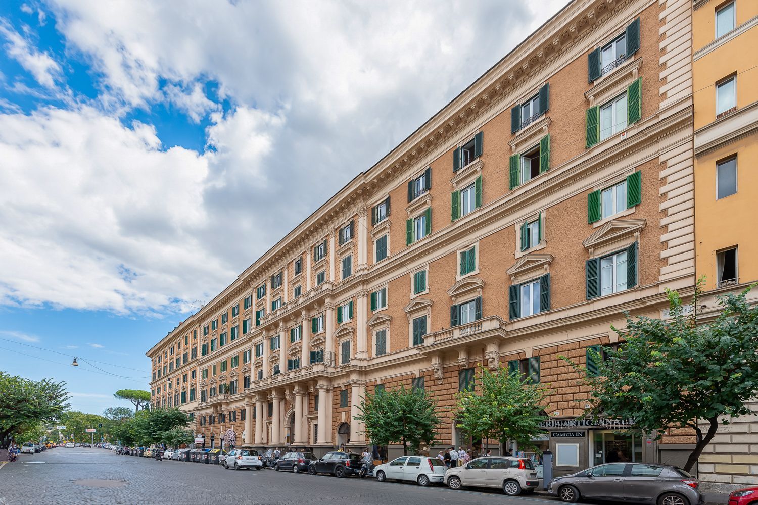 A multi-story building with a classical architectural style, featuring a row of windows with green shutters, a central entrance with a portico, and a street view with parked cars and pedestrians.