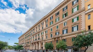 A multi-story building with a classical architectural style, featuring a row of windows with green shutters, a central entrance with a portico, and a street view with parked cars and pedestrians.