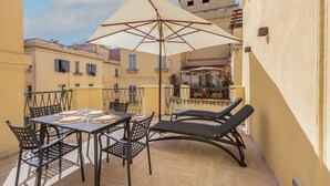 A balcony with a table set for two, two lounge chairs, and a large umbrella providing shade.