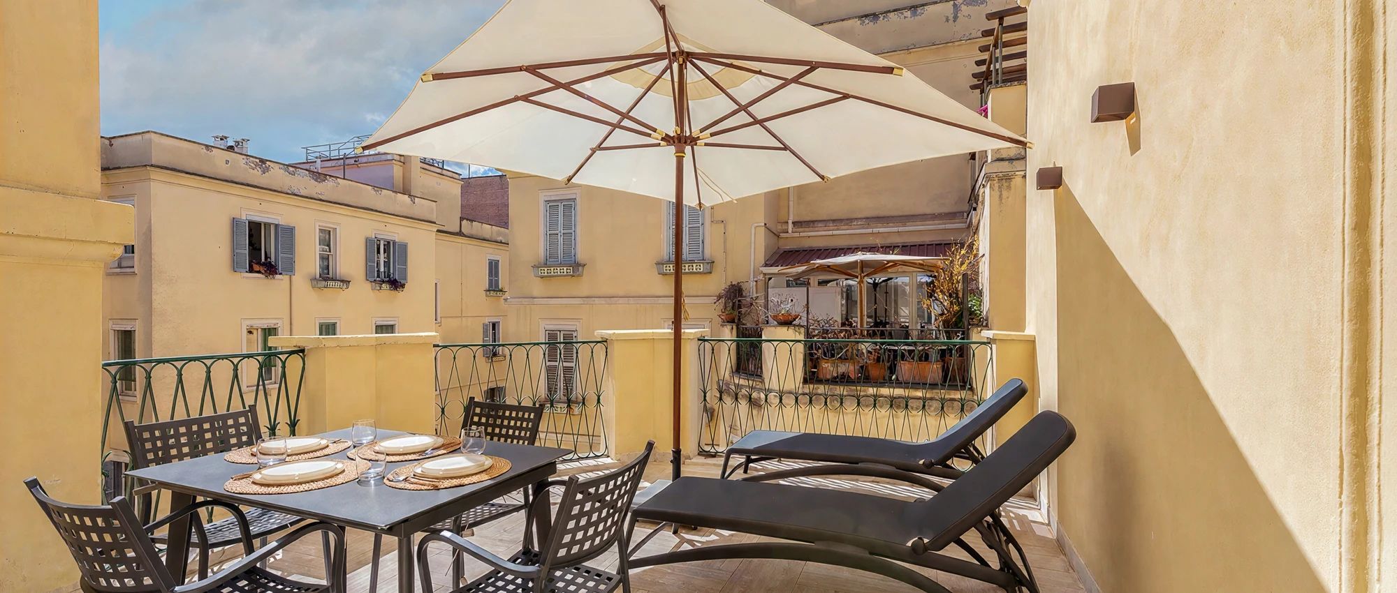 En balkon med et spisebord, en liggestol og en stor parasol, der giver skygge.