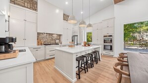 A modern kitchen with white cabinetry, a large island with bar stools, a stone accent wall, and a view of the outdoors.