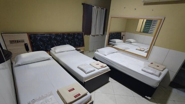 Hotelkamer met drie enkele bedden, elk voorzien van een kussen en dekbed. Aan de muur hangt een spiegel, er is een raam met gordijnen en een airconditioning boven de bedden.