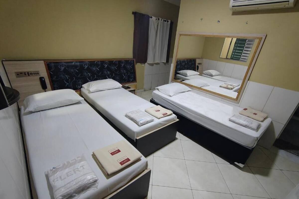 Hotelkamer met drie enkele bedden, elk voorzien van een kussen en dekbed. Aan de muur hangt een spiegel, er is een raam met gordijnen en een airconditioning boven de bedden.