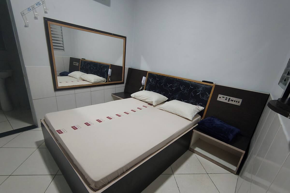 Een hotelkamer met een bed, nachtkastje, spiegel en bedieningspaneel aan de muur.