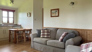 Una sala de estar acogedora con un sofá gris, una mesa de comedor de madera y sillas. Se ve una cocina al fondo con una ventana y un ventilador de techo.