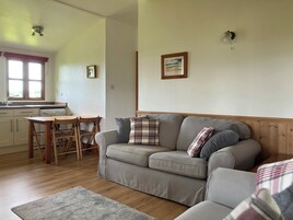 Una sala de estar acogedora con un sofá gris, una mesa de comedor de madera y sillas. Se ve una cocina al fondo con una ventana y un ventilador de techo.