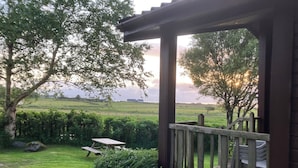 Un patio techado con terraza de madera, una mesa de picnic y vista a un campo cubierto de césped, árboles y un edificio lejano.
