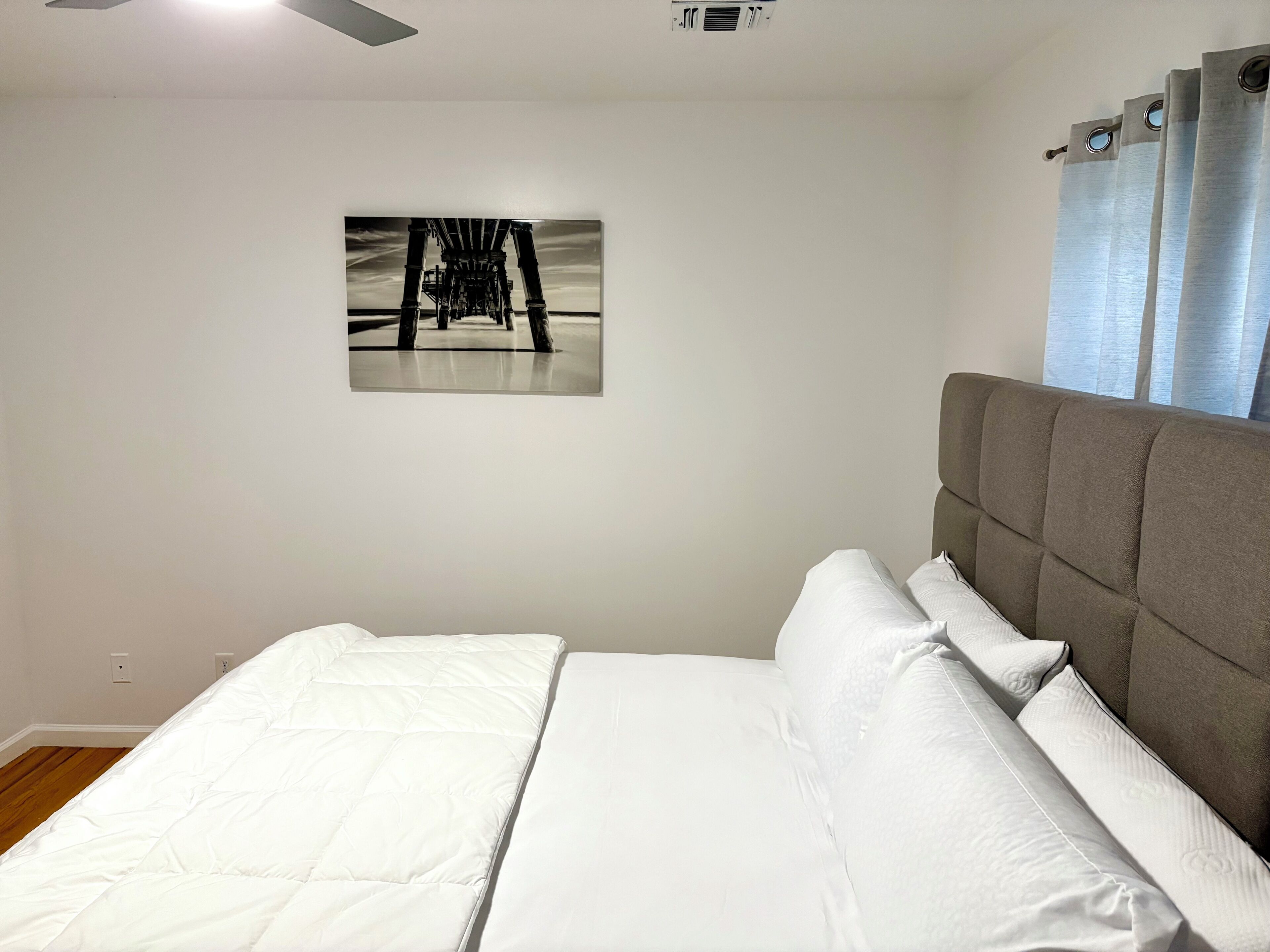 A bedroom with a bed, a grey headboard, a ceiling fan, a wall-mounted air conditioning unit, and a framed picture on the wall.