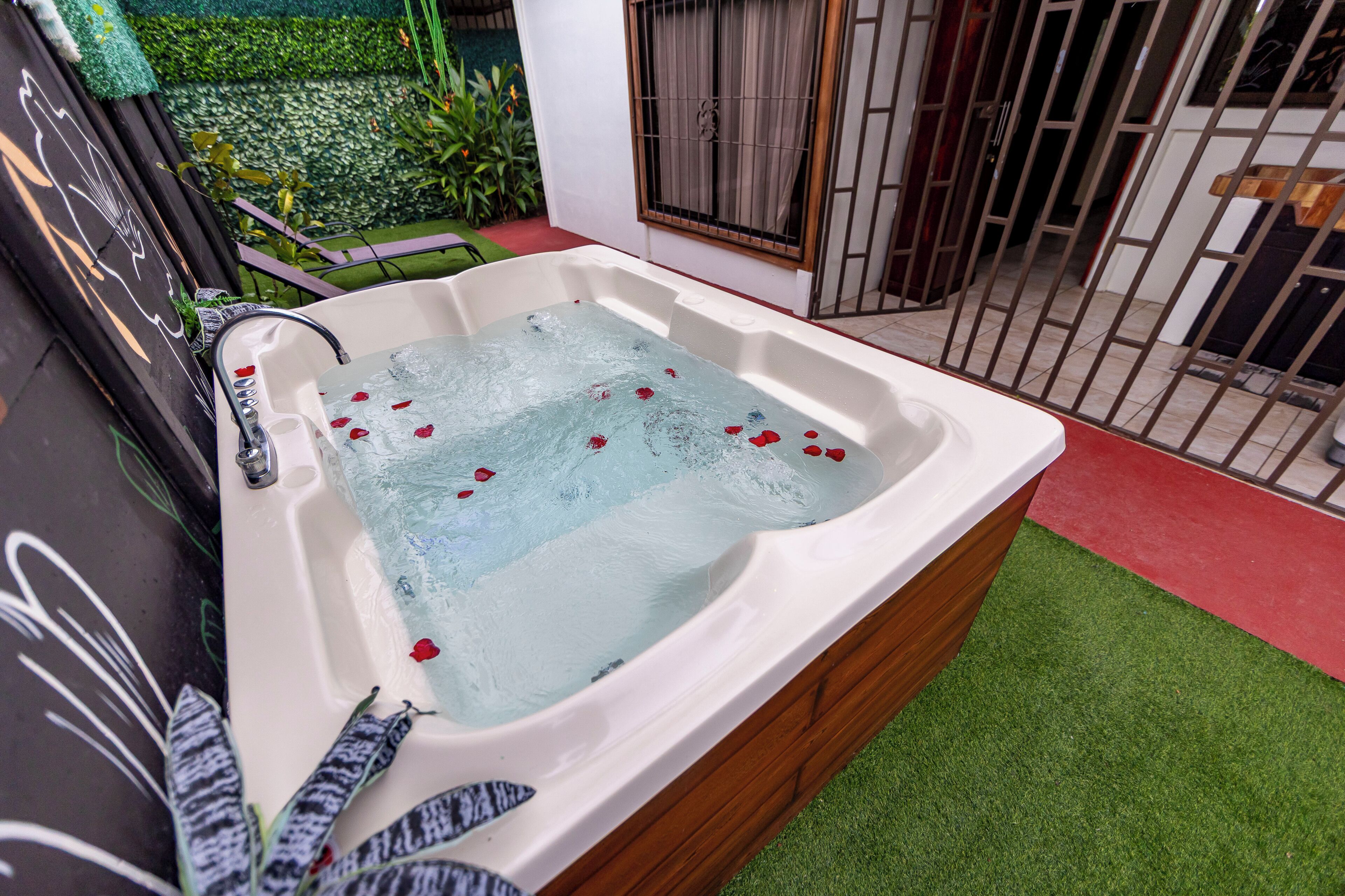 A hot tub with water and rose petals, surrounded by a wooden deck and a green artificial turf area.