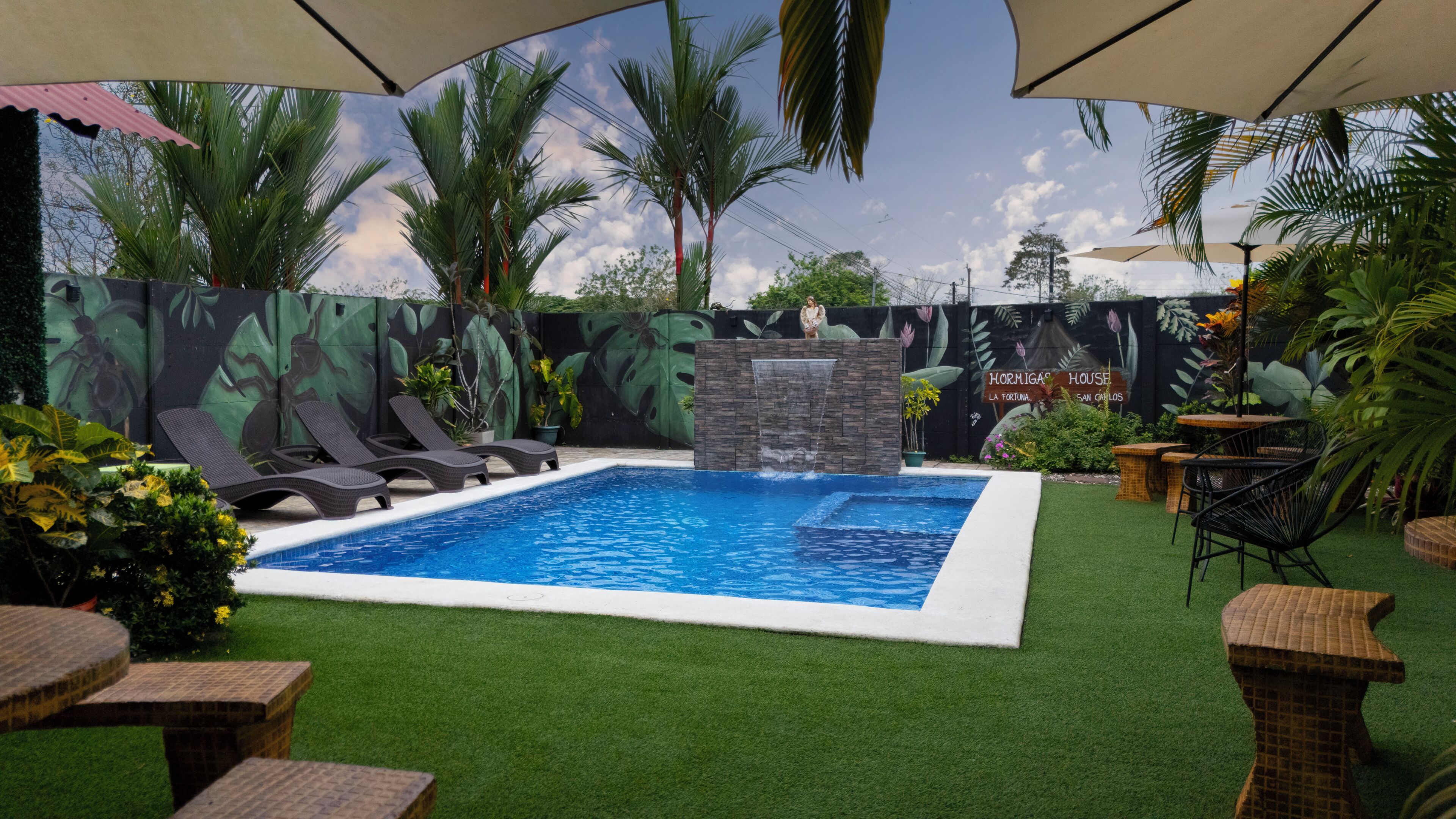 A swimming pool with a waterfall feature, surrounded by artificial grass, palm trees, and lounge chairs.