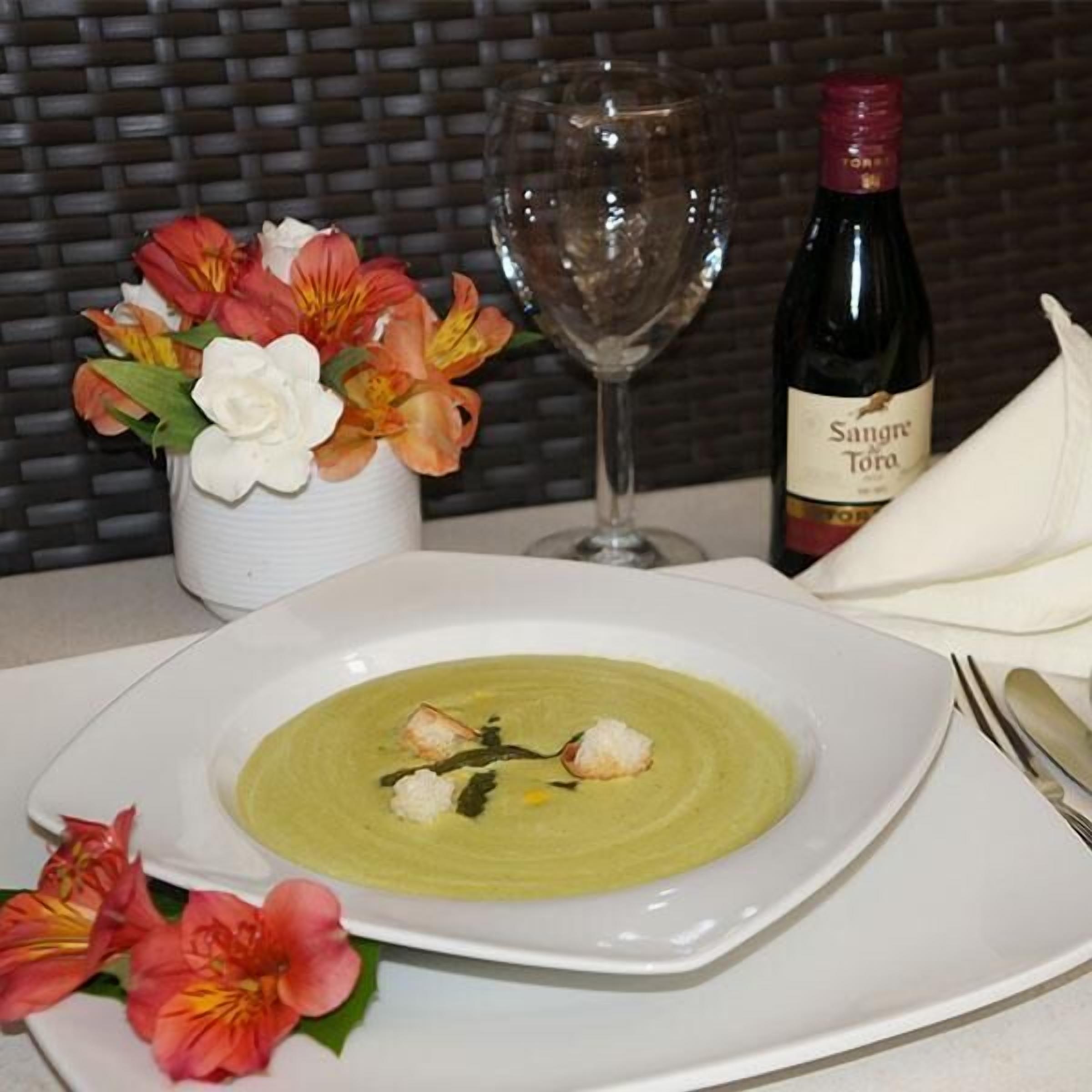 A dining table with a bowl of soup, a wine bottle, a glass, and a vase of flowers.