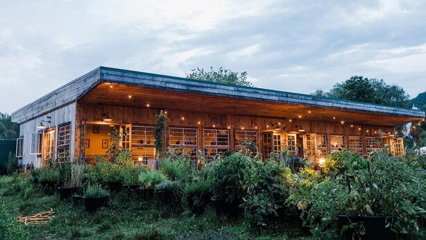 Un bâtiment rustique en bois, doté de nombreuses fenêtres et d’un porche abrité, entouré d’une végétation luxuriante.