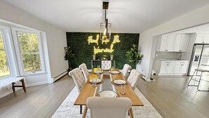 Un espace repas moderne, avec une longue table en bois dressée pour six personnes, entourée de chaises à revêtement blanc. Un néon au-dessus de la table affiche l’inscription : « Trouvez du plaisir dans votre travail. » La pièce est dotée d’un mur vert à motifs texturés, de grandes fenêtres et d’un coin cuisine avec des armoires blanches et un réfrigérateur.
