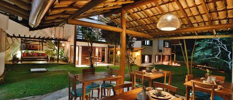 An outdoor dining area with wooden tables and chairs, a covered seating area with a woven ceiling, and a well-maintained lawn.