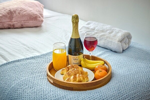 A breakfast tray with croissants, bananas, oranges, a bottle of bubbly, a glass of orange juice, and a glass of red wine on a bed with a blue blanket and a pink pillow.
