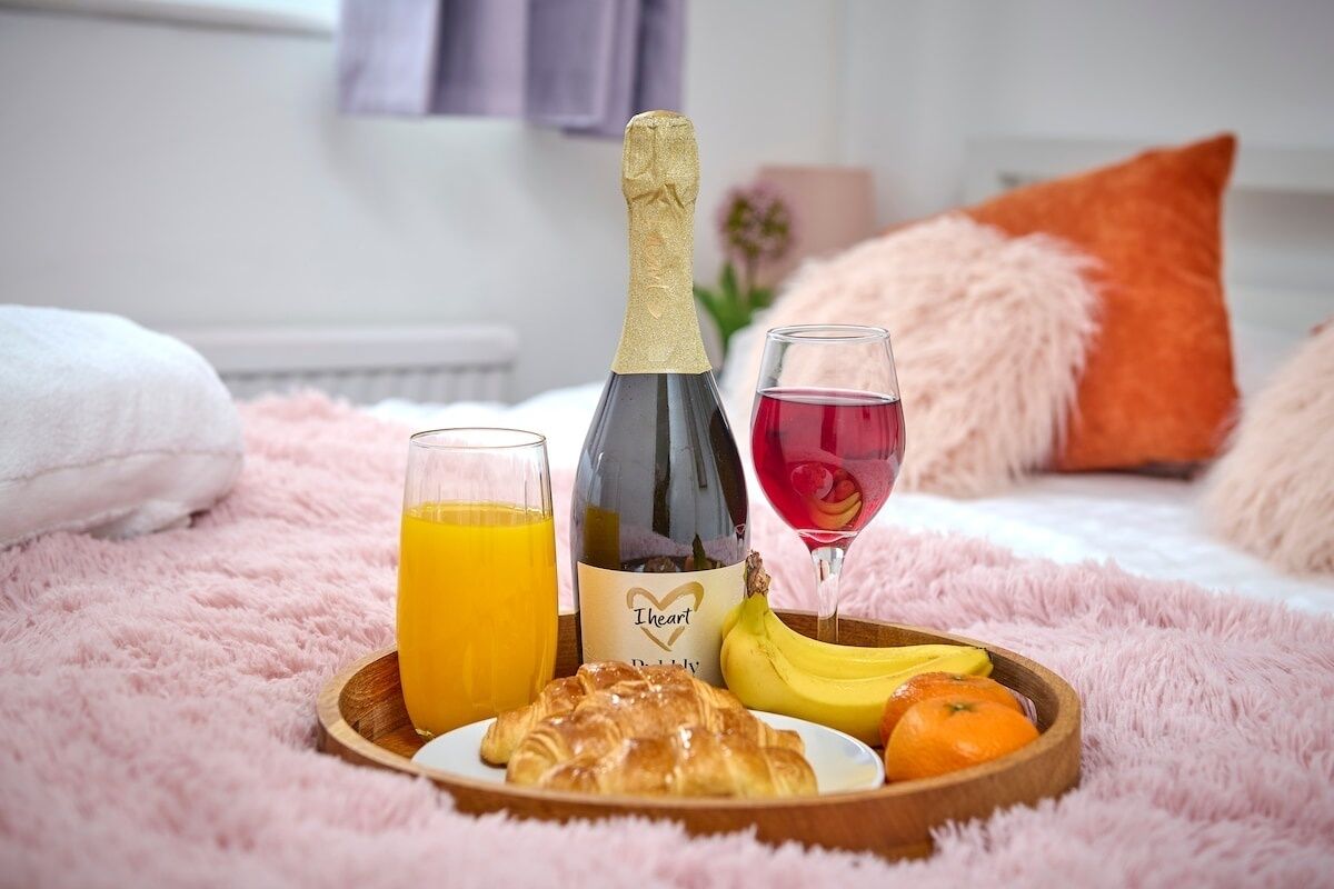 A breakfast setup on a bed with a bottle of champagne, a glass of red wine, a glass of orange juice, a croissant, a banana, and an orange.