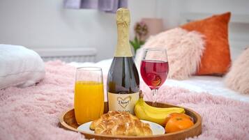 A breakfast setup on a bed with a bottle of champagne, a glass of red wine, a glass of orange juice, a croissant, a banana, and an orange.