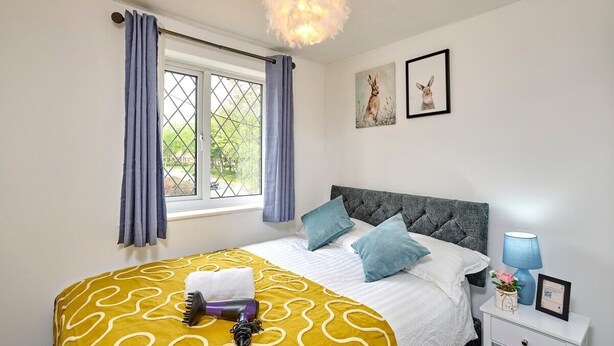 A neatly made bed with a patterned bedspread, a grey upholstered headboard, and a bedside table with a lamp and a framed picture. - Home in Milton Keynes (Great Holm)