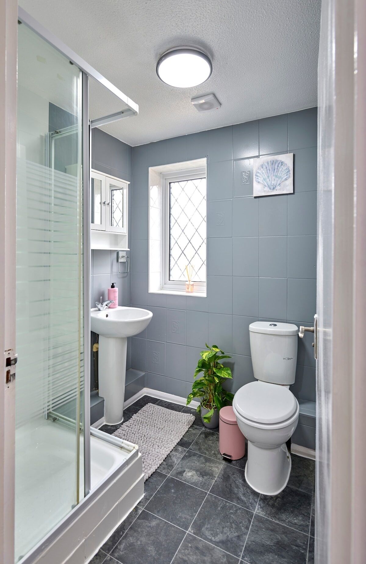A bathroom with a glass shower enclosure, a pedestal sink, a toilet, and a window with a decorative item.