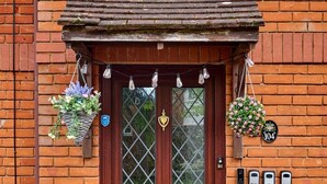 A wooden front door with a glass panel, a brass handle, and a small brass knocker. There are two hanging flower baskets, a doormat with a message, and a house number plaque. - Home in Milton Keynes (Great Holm)