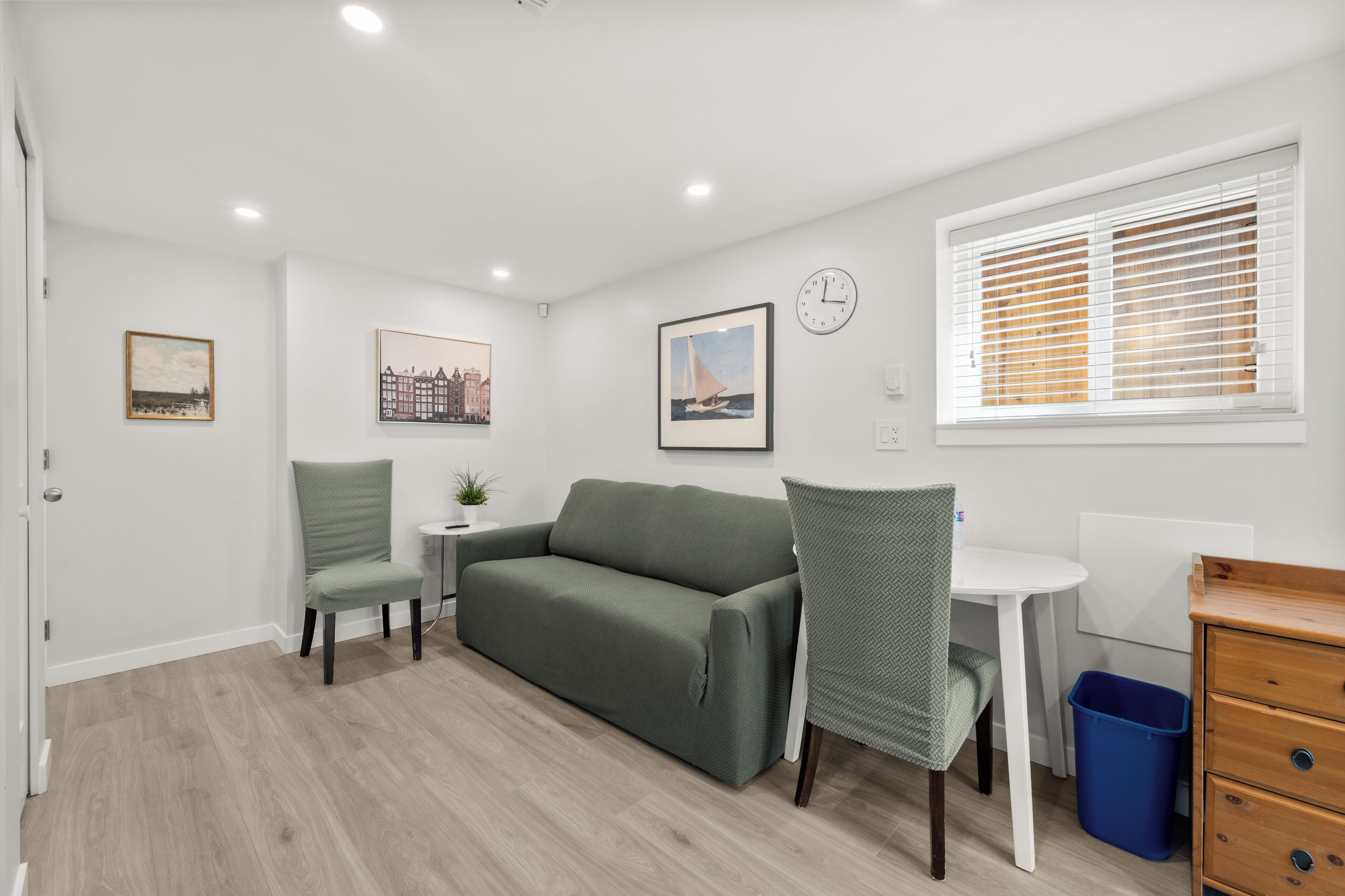 A room with a green sofa, two chairs, a small table, a clock, and a window with blinds.
