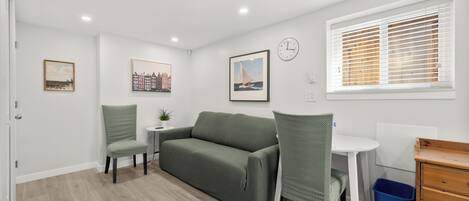 A room with a green sofa, two chairs, a small table, a clock, and a window with blinds.