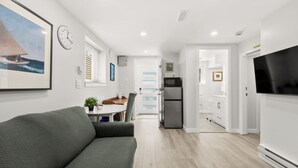 A modern living room with a sofa, a flat-screen TV, a dining area with a table and chairs, and a kitchen with a refrigerator and microwave.