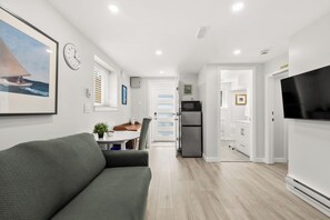 A modern living room with a sofa, a flat-screen TV, a dining area with a table and chairs, and a kitchen with a refrigerator and microwave.