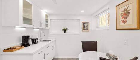 A modern kitchen with white cabinetry, a coffee maker, and a dining area with a white table and brown chairs.