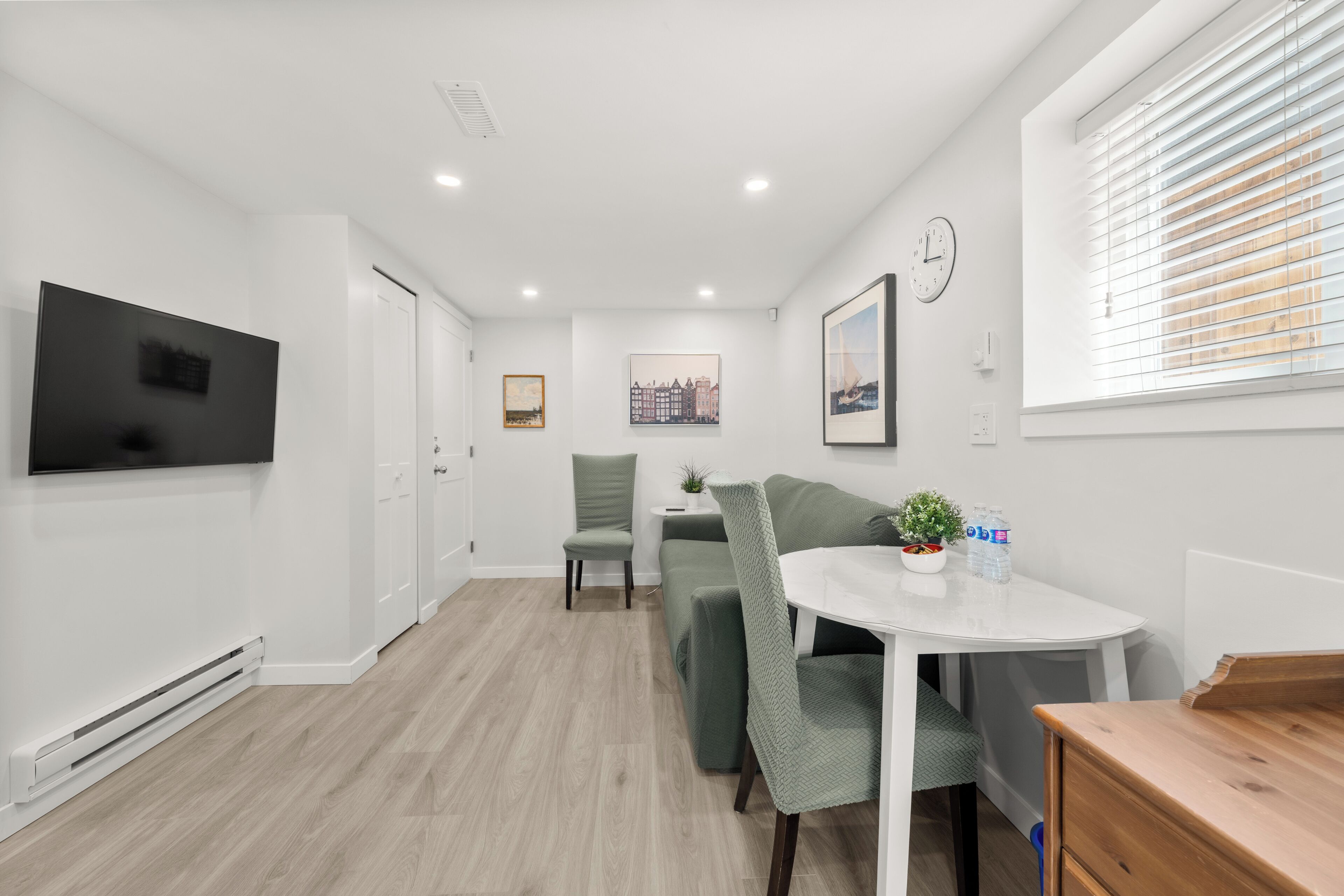 A modern room with a flat-screen TV, a sofa, a small table with a plant, a clock, and a window with blinds.