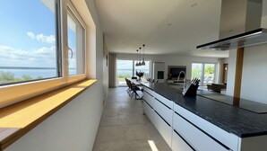 A modern kitchen with a large island, a dining area with chairs, and a view of the sea through large windows.