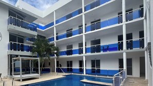 Ein mehrstöckiges Hotel mit Schwimmbad, Balkonen mit blauen Geländern und einer Palme.