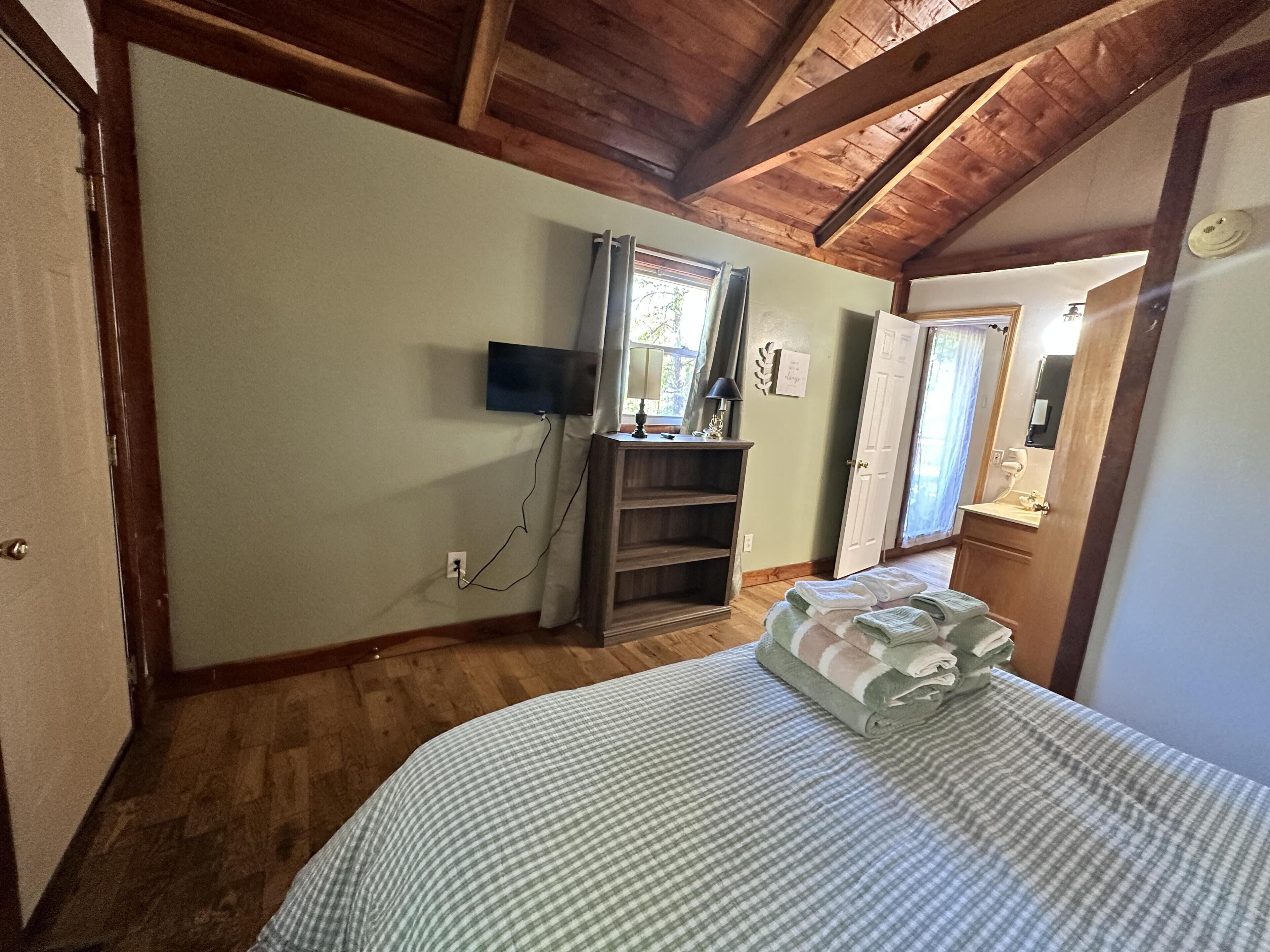 A bedroom with a bed, a TV mounted on the wall, a wooden ceiling, and a door leading to another room.
