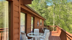 A wooden deck with a glass table and white chairs, surrounded by a wooden railing, overlooking a forested area.
