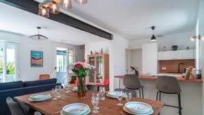 A modern dining area with a wooden table set for six, surrounded by chairs. A vase with flowers is on the table, and there is a kitchen area in the background.