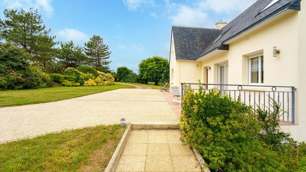 A residential property with a paved driveway leading to a house with a dark roof and light walls.