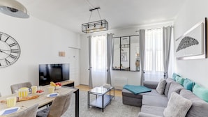 A modern living room with a flat-screen TV, a dining area with striped cups and a pizza, a gray sofa with teal cushions, and a large wall clock. - Jungle d'orient - Proche centre ville Montluçon (Montlucon)