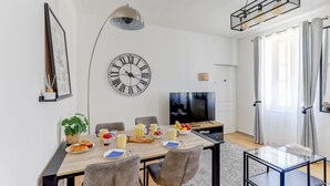 A modern living room with a dining area, featuring a wooden table, chairs, a large wall clock, a flat-screen TV, and a hanging light fixture. - Jungle d'orient - Proche centre ville Montluçon (Montlucon)