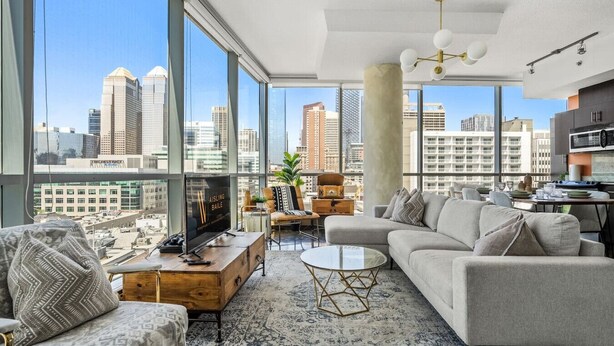 A modern living room with a large window, a flat-screen TV, a sofa, a coffee table, and a dining area with a kitchen in the background.