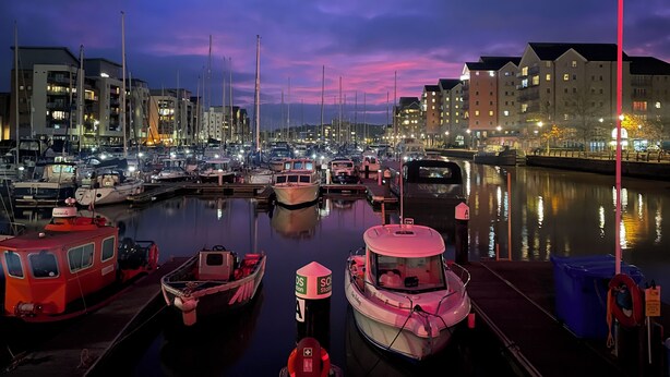 A marina at dusk with boats docked, buildings in the background, and a calm water surface reflecting the lights. - Stylish Marina Apartment with Stunning Views (Portishead)