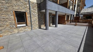 A modern building with stone and concrete exterior walls, a balcony with wooden railings, and a tiled patio area.