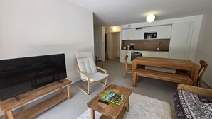 A modern living room with a flat-screen TV, a comfortable armchair, a wooden coffee table with a magazine, and a dining table with chairs.