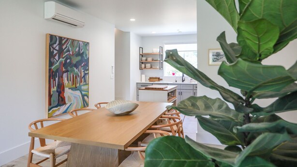 Un comedor moderno con una mesa y sillas de madera, un cuadro abstracto grande en la pared y una cocina al fondo.