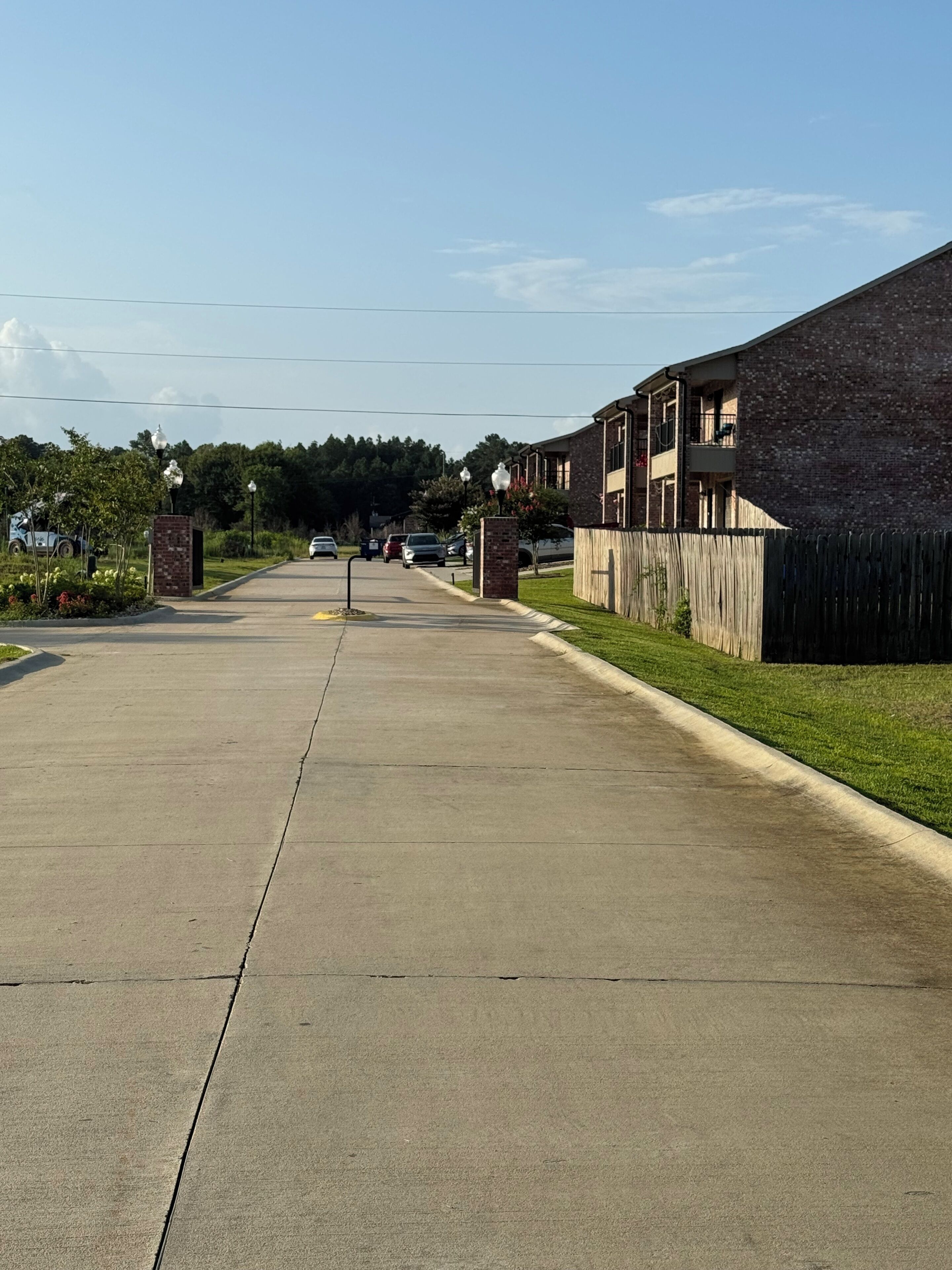 A long, straight driveway leading to a residential area with brick buildings and a wooden fence.