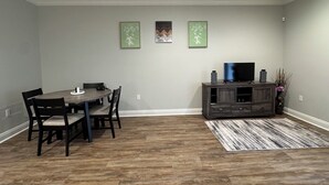A dining area with a wooden table and chairs, a dark wooden cabinet with a flat-screen TV, and two framed artworks on the wall.